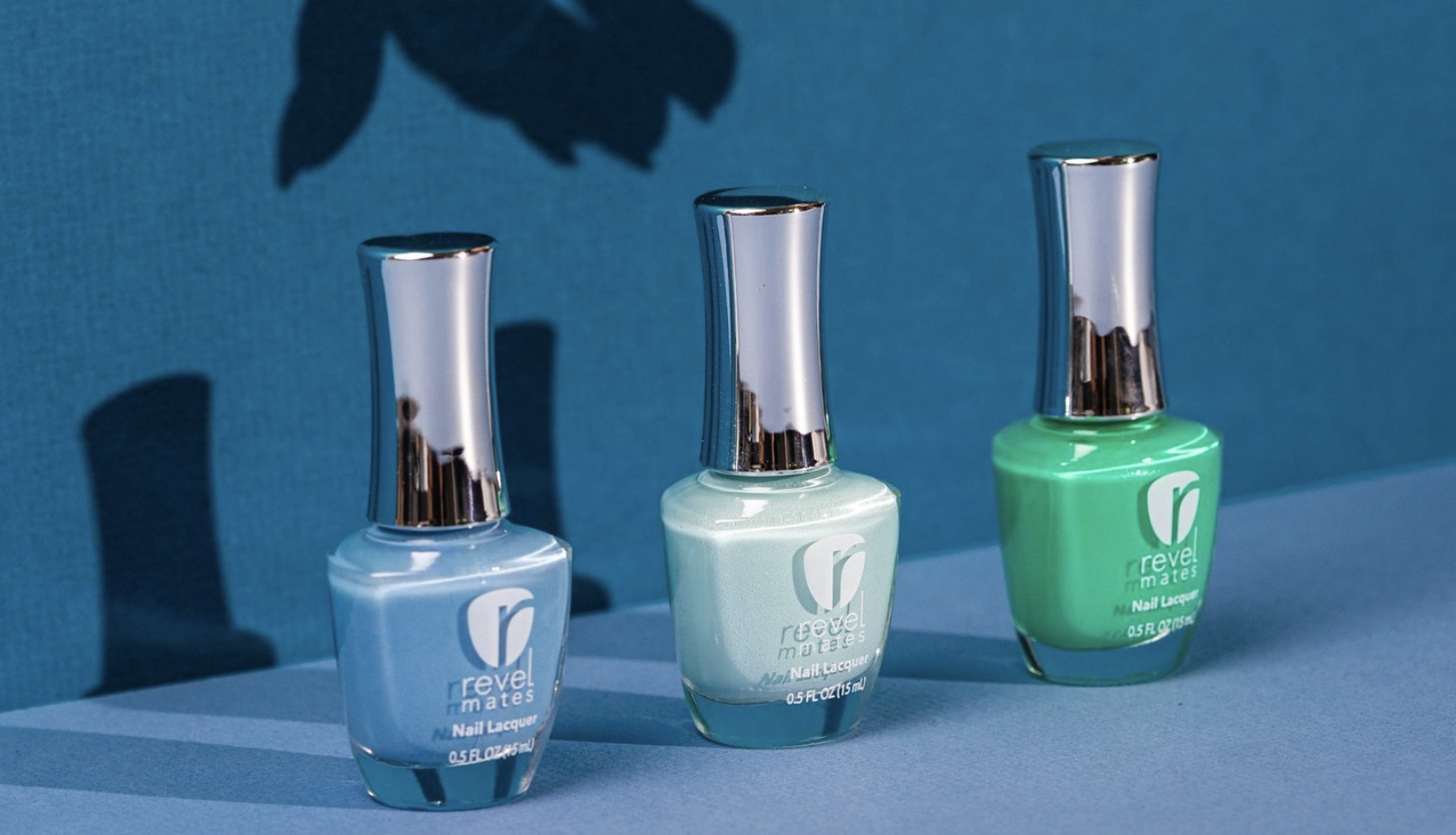 Three bottles of Revel Nail nail polish in blue, mint, and green. The background behind them in blue and they're placed in a